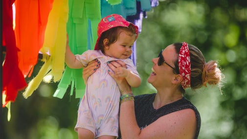 A woman holding a babay wearing a sun hat with rainbow of ribbons in the background